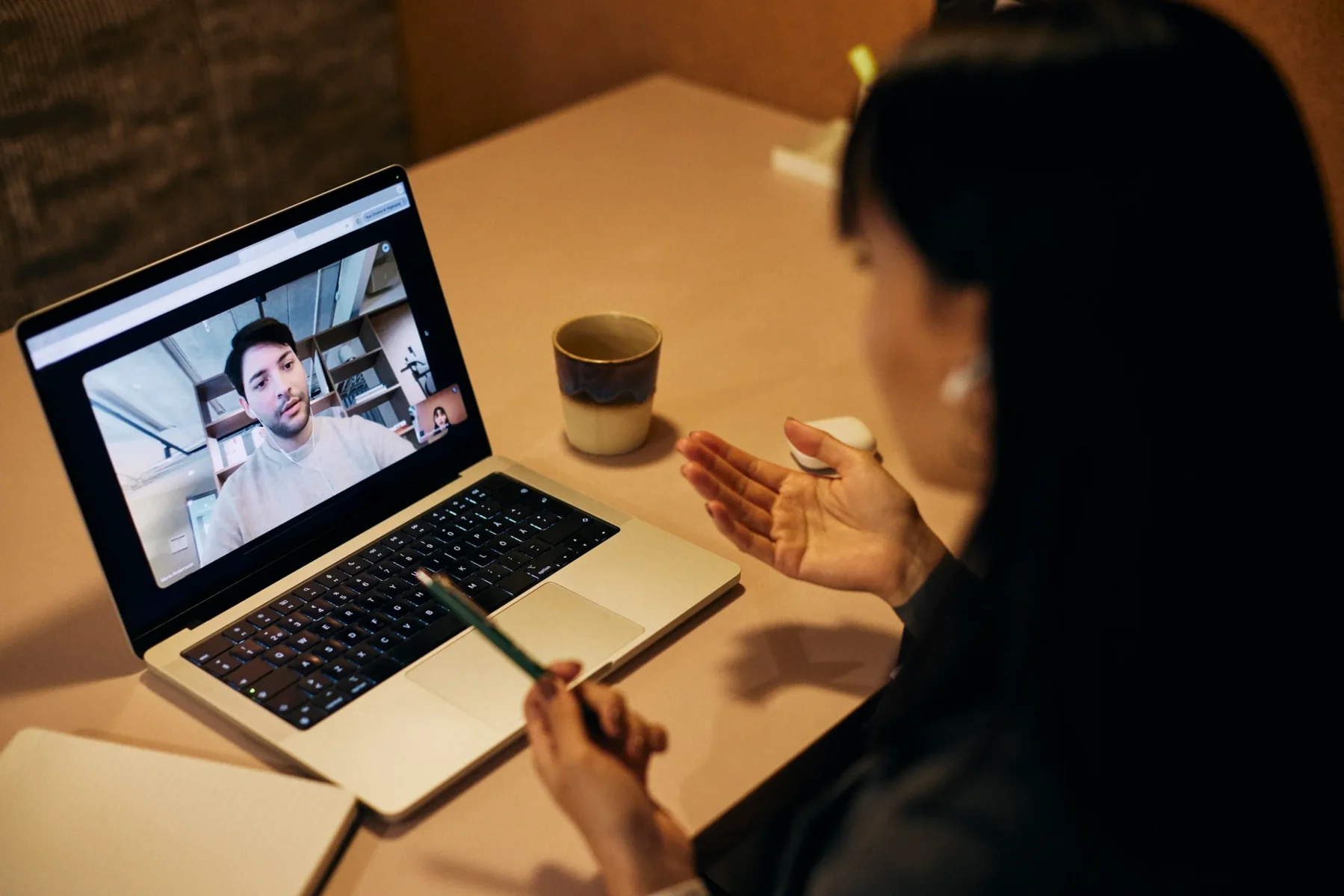 En person deltar i ett videosamtal på en bärbar dator och talar med en man som syns på skärmen. På bordet ligger en anteckningsbok, en kopp och ett par hörlurar i etui.