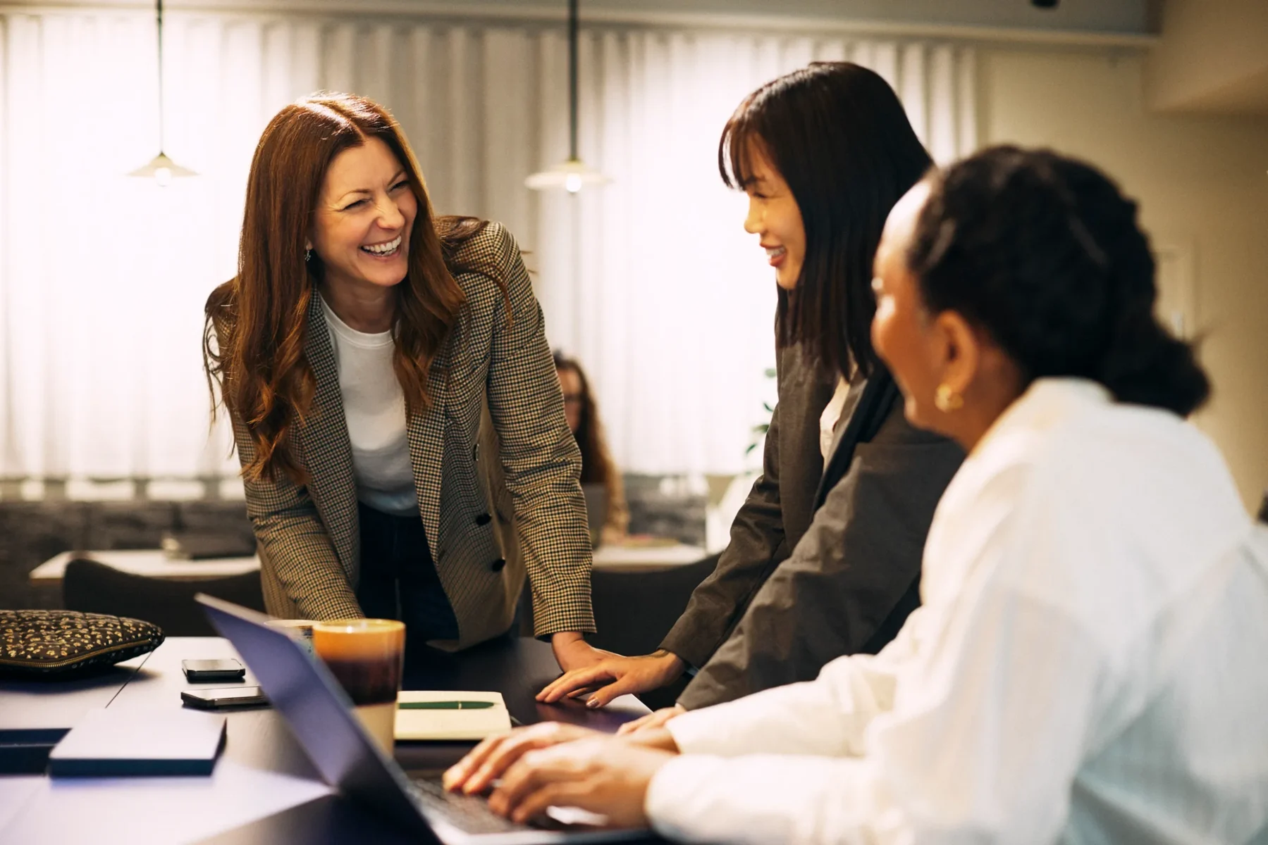 Tre personer står och sitter runt ett bord i ett kontor och skrattar tillsammans. En laptop står öppen framför dem och andra arbetsmaterial ligger utspridda på bordet. Omgivningen är varmt upplyst och avslappnad.