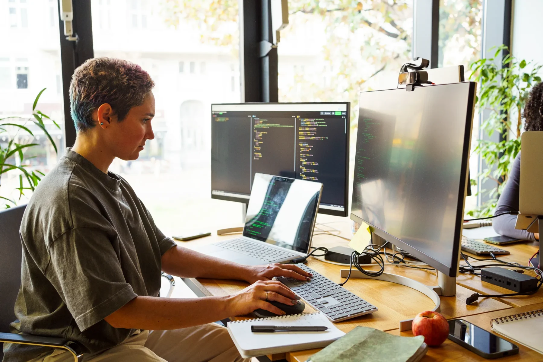 En person sitter vid ett skrivbord med flera skärmar och arbetar med kod på datorn. Framför sig har personen en laptop, två stora bildskärmar, tangentbord, mus samt ett anteckningsblock. Ljuset från stora fönster lyser in i det moderna kontoret.