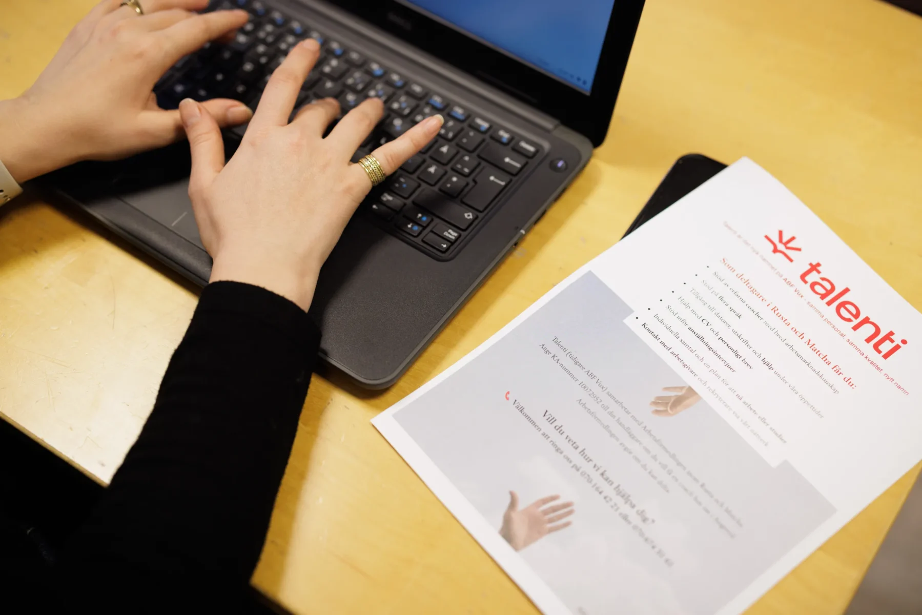 Närbild av händer som skriver på en bärbar dator bredvid ett informationsblad från Talenti som ligger på ett träbord.