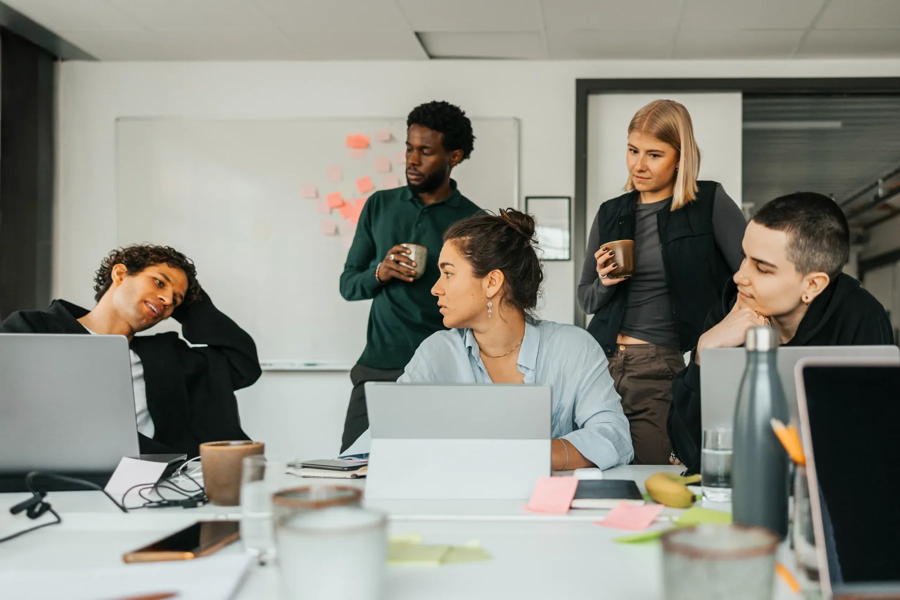 En grupp unga vuxna arbetar tillsammans runt ett bord med laptops, anteckningar och kaffekoppar, medan några står och håller muggar och pratar framför en whiteboard med post it-lappar.