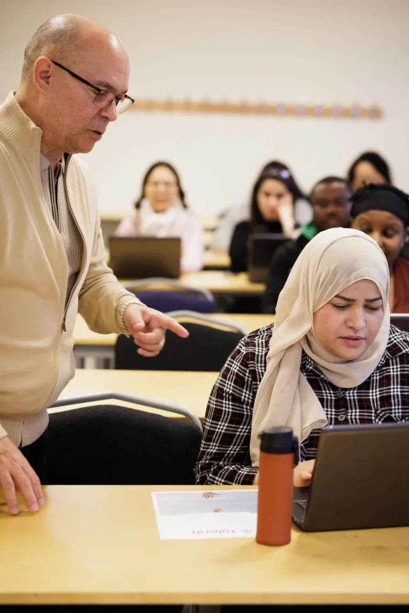 En lärare lutar sig fram och ger stöd till en kvinna som arbetar vid en laptop i ett klassrum, medan andra deltagare sitter och jobbar i bakgrunden.