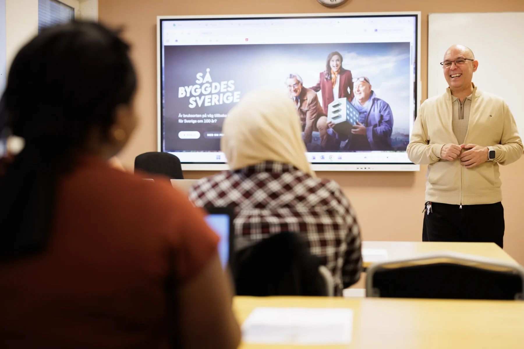 En lärare står leende framför en skärm som visar en presentation med texten Så byggdes Sverige, medan deltagare sitter vid bänkar och lyssnar.