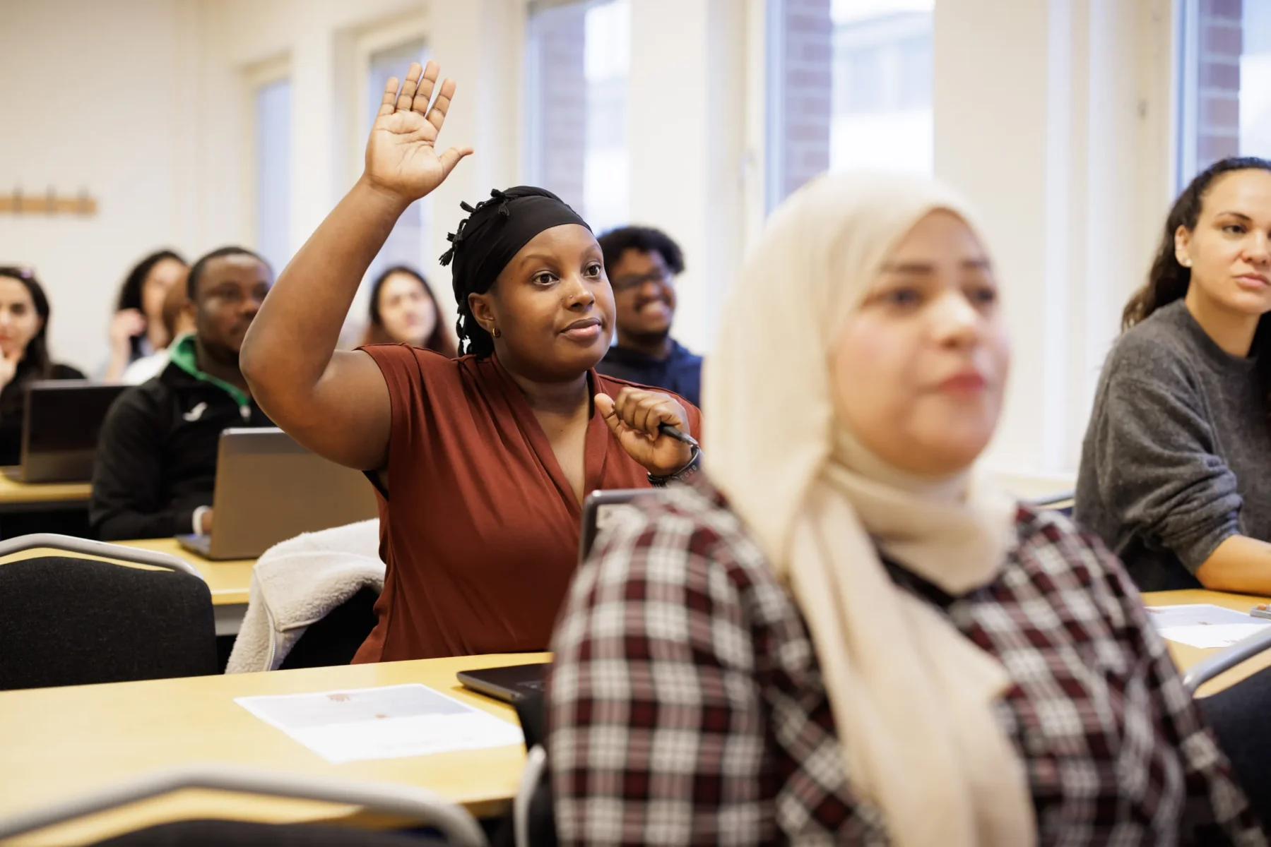 En kvinna i ett klassrum räcker upp handen för att ställa en fråga, medan andra deltagare sitter vid sina bänkar och lyssnar.