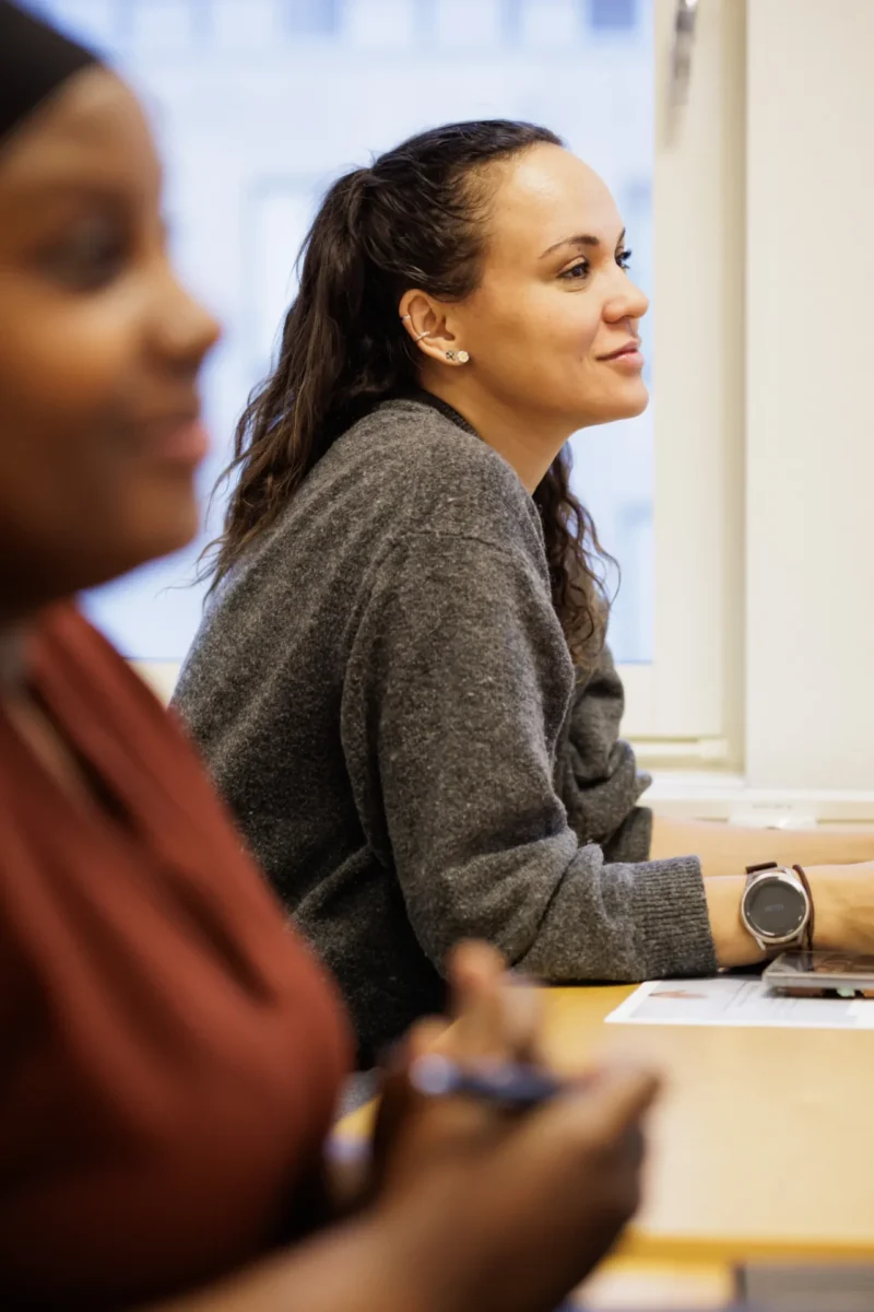 En kvinna sitter vid en bänk i ett klassrum och lyssnar uppmärksamt, med en annan deltagare suddigt i förgrunden.