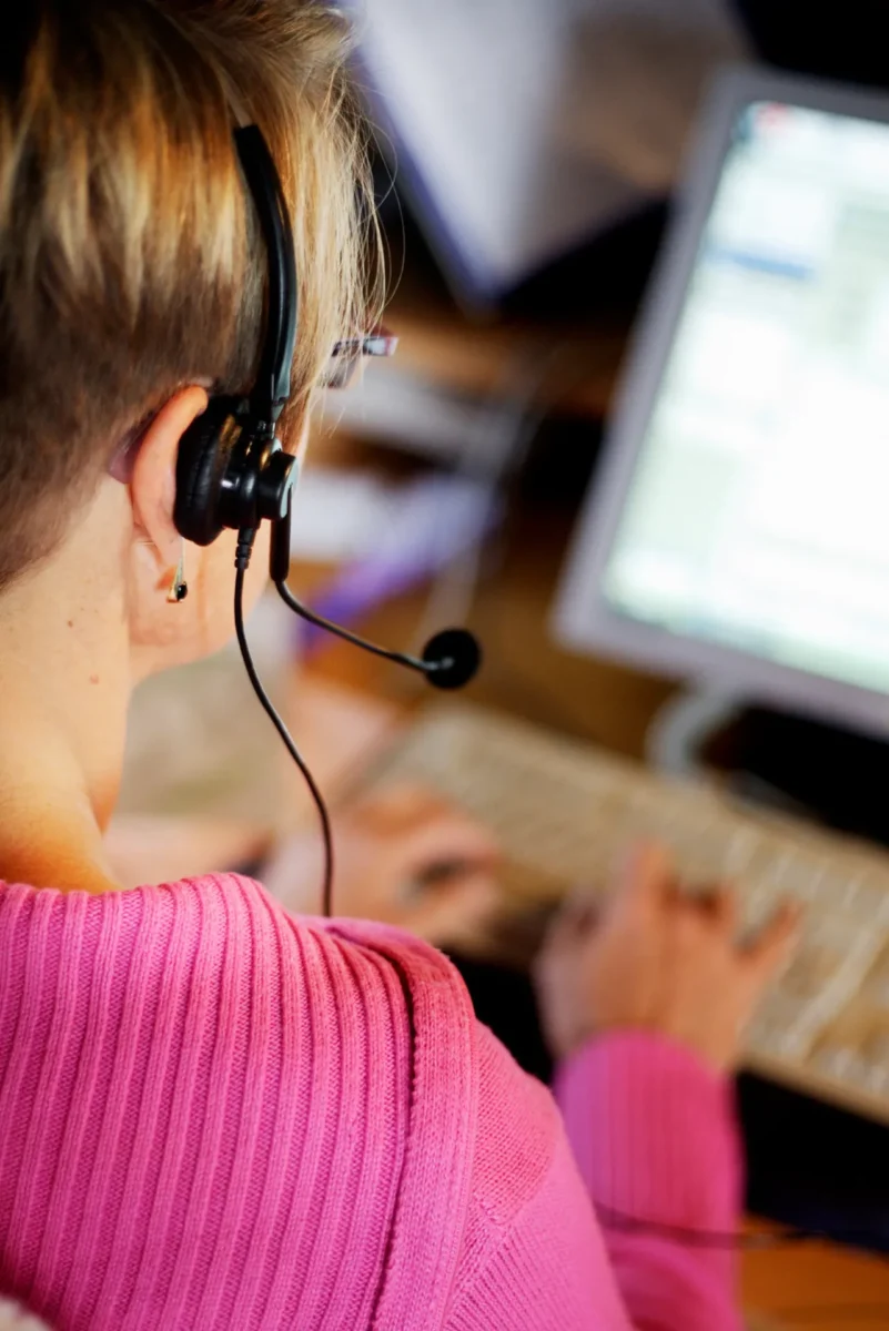 En person med headset sitter vid en dator och skriver på ett tangentbord. Bilden är tagen bakifrån och fokus ligger på headsetet och arbetssituationen.